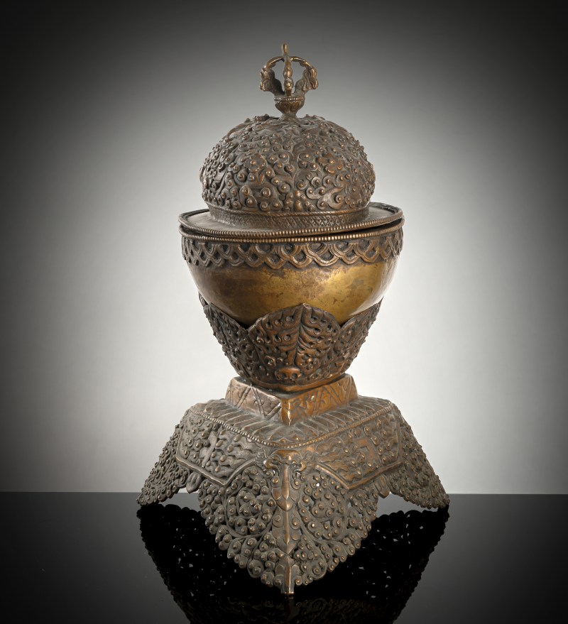 A COPPER EMBOSSED KAPALA WITH COVER ON STAND
